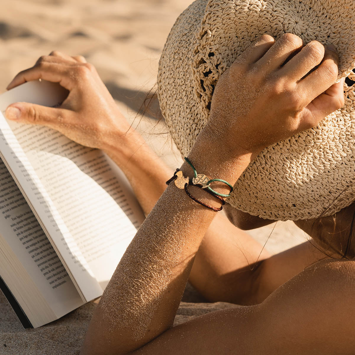 Woman wearing a straw hat and bracelets reading a book on the beach. Sunlit scene with sand-covered arms enhancing the summertime leisure vibe.